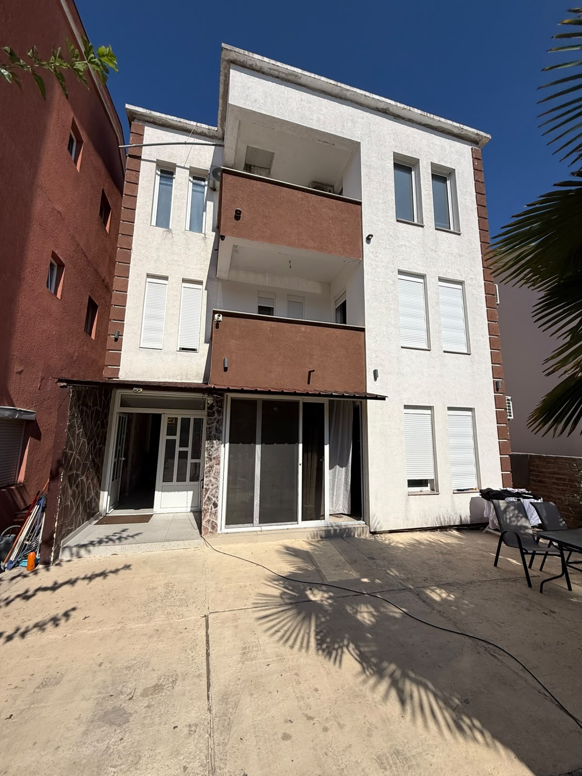 Modern three-story residential building for sale in Ulcinj, Montenegro, featuring elegant white and brown exterior finishes. The property offers spacious balconies on the second and third floors, expansive windows for abundant natural light, and a stylish glass patio door connecting to a charming outdoor seating area on a paved terrace. Located under clear blue skies in a desirable Montenegrin neighborhood, this listing is an exceptional investment opportunity in the fast-growing Ulcinj real estate market. Discover more exclusive properties at montenegro-listings.me—a project by ekosphere doo in Ulcinj—connecting you to premium Montenegro investments.