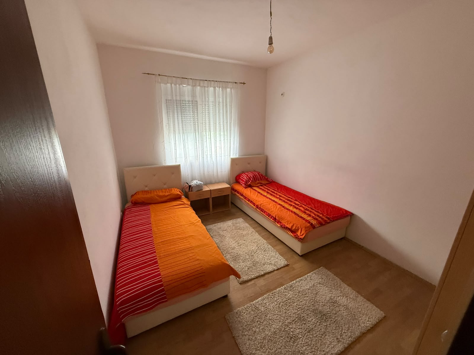 Discover a cozy and inviting small bedroom in Ulcinj, Montenegro, featuring two comfortable single beds with vibrant orange-red bedding, ideal for guests or family stays. The room is filled with natural light through a large window adorned with a sheer curtain, enhancing the serene atmosphere. A stylish wooden nightstand sits elegantly between the beds for added convenience, while two soft beige rugs complement the polished wooden floor. This thoughtfully designed space offers both comfort and functionality—an excellent real estate investment opportunity highlighted exclusively on montenegro-listings.me, a project of ekosphere doo in Ulcinj. Perfect for buyers seeking property value and rental potential in Montenegro’s thriving coastal market.